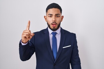 Young hispanic man wearing business suit and tie pointing finger up with successful idea. exited and happy. number one.