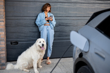 Woman uses her phone leaning against the garage door of her house, a white dog by her side. An...