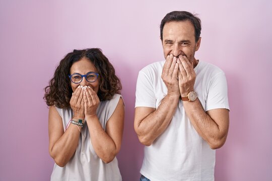 Middle age hispanic couple together over pink background laughing and embarrassed giggle covering mouth with hands, gossip and scandal concept