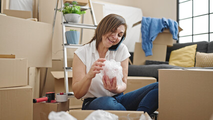 Fototapeta premium Middle age hispanic woman unpacking cardboard box speaking on the phone at new home