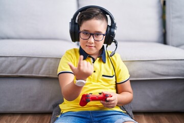 Young hispanic kid playing video game holding controller wearing headphones beckoning come here gesture with hand inviting welcoming happy and smiling