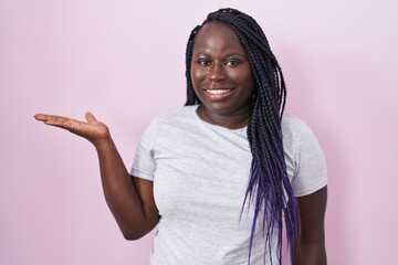 Young african woman standing over pink background smiling cheerful presenting and pointing with palm of hand looking at the camera.