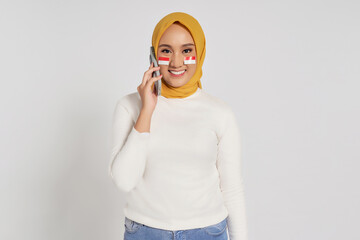 Smiling happy young Asian Muslim woman wearing a hijab talking on mobile phone and looking at camera isolated over white background. Celebrate Indonesian independence day on 17 August