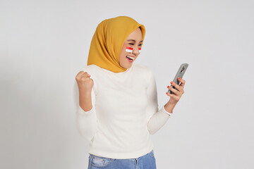 Excited young Asian Muslim woman wearing a hijab using smartphone and celebrating Indonesian...