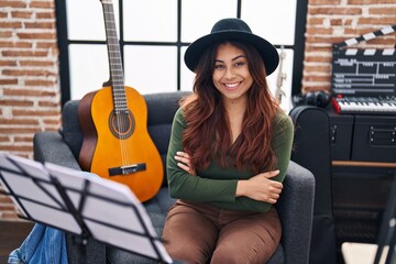 Young hispanic woman musician sitting with arms crossed gesture at music studio