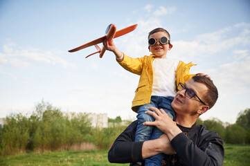 Boy is sitting on man's shoulders. Father with his little son playing with toy plane on the field - obrazy, fototapety, plakaty