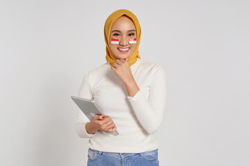 Smiling young Asian Muslim woman wearing a hijab holding digital tablet and hand touching her chin isolated over white background. Celebrate Indonesian independence day on 17 August