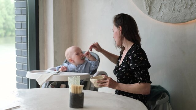 Mother feeds a little boy with a spoon. Mother giving food to her adorable baby at home