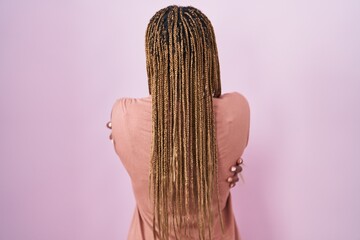 African american woman with braided hair standing over pink background hugging oneself happy and positive from backwards. self love and self care