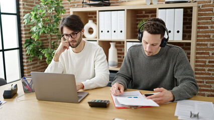 Two men business partners using laptop and headphones writing on document working at office