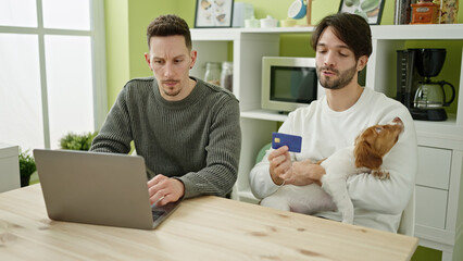 Obraz premium Two men couple shopping with laptop and credit card sitting on table with dog at dinning room