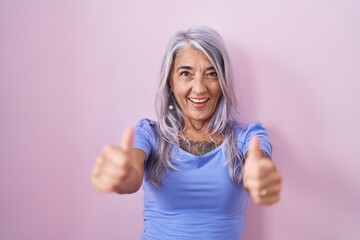 Middle age woman with tattoos standing over pink background approving doing positive gesture with hand, thumbs up smiling and happy for success. winner gesture.