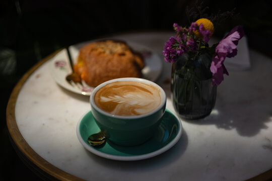 A foamy cappuccino is served in a green ceramic cup on a saucer, with a croissant in the background. It is set on a marble table with a table setting of purple flowers. it is lit in hard light.