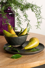 Three green pears lie in a ceramic plate on the table by a purple vase with green twigs.