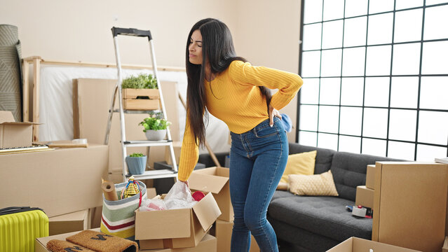 Young beautiful hispanic woman holding cardboard box suffering for backache at new home