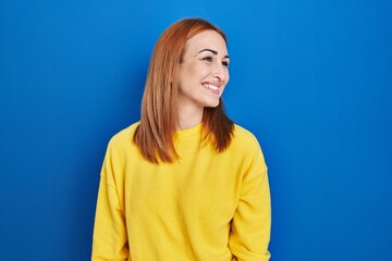 Young woman standing over blue background looking away to side with smile on face, natural...