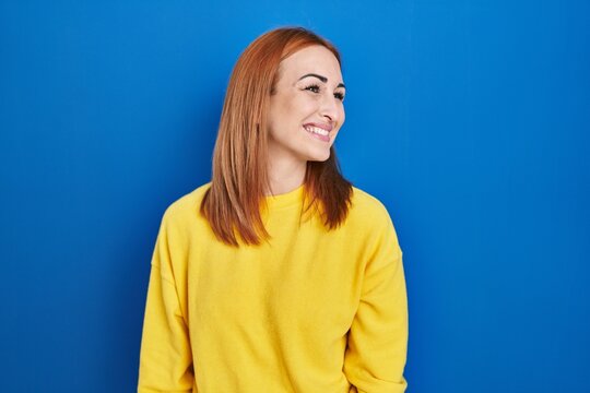 Young woman standing over blue background looking away to side with smile on face, natural expression. laughing confident.