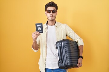 Young hispanic man holding suitcase going on summer vacation holding italian passport making fish...
