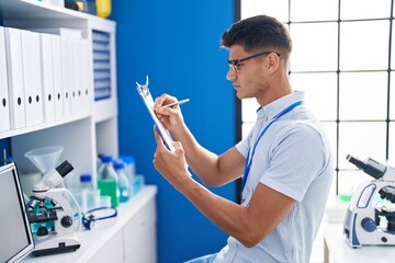 Young hispanic man scientist writing on document at laboratory