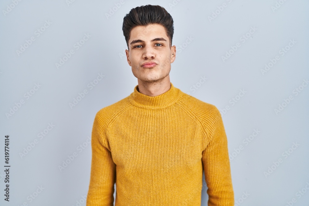 Wall mural Young hispanic man standing over blue background looking at the camera blowing a kiss on air being lovely and sexy. love expression.