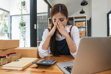 Small online business owner Asian woman wearing apron stressed headache taking notes with parcel box at home