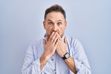 Middle age caucasian man standing over blue background shocked covering mouth with hands for...