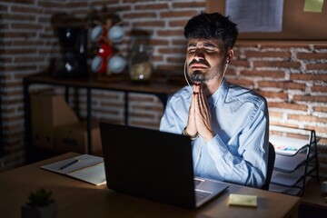 Plakat Young hispanic man with beard working at the office at night begging and praying with hands together with hope expression on face very emotional and worried. begging.