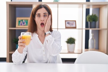 Brunette woman drinking glass of orange juice afraid and shocked, surprise and amazed expression with hands on face