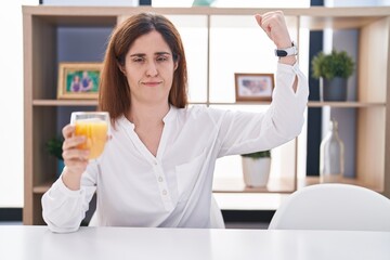 Brunette woman drinking glass of orange juice strong person showing arm muscle, confident and proud of power