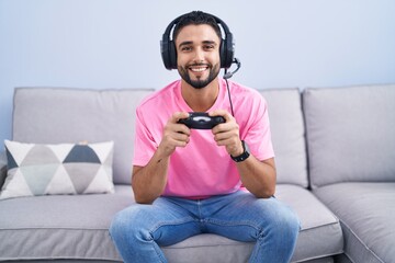Hispanic young man playing video game holding controller sitting on the sofa with a happy and cool...