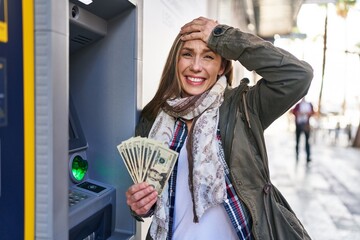 Young blonde woman holding dollars banknotes from atm machine stressed and frustrated with hand on...
