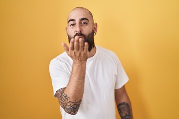 Young hispanic man with beard and tattoos standing over yellow background looking at the camera blowing a kiss with hand on air being lovely and sexy. love expression.