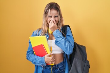 Young blonde woman wearing student backpack and holding books smelling something stinky and disgusting, intolerable smell, holding breath with fingers on nose. bad smell