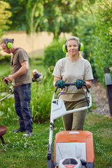 Young Female gardener using a lawn mower while her colleague or bosses is trimming the garden in the background