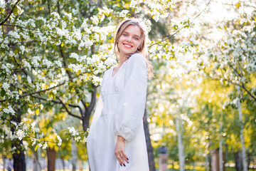 Portrait of a young woman in a white dress against the backdrop of blooming apple trees. The girl poses against the background of flowers in the spring park.