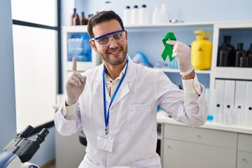 Young hispanic man with beard working at scientist laboratory holding green ribbon surprised with an idea or question pointing finger with happy face, number one