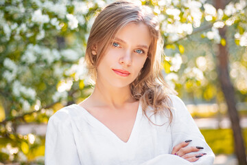 Portrait of a young woman in a white dress against the backdrop of blooming apple trees. The girl poses against the background of flowers in the spring park.