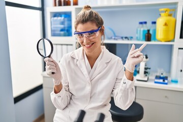 Young woman working at scientist laboratory holding magnifying glass smiling happy pointing with hand and finger to the side