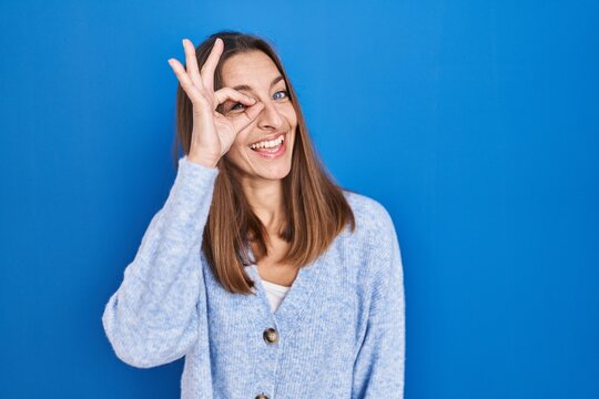 Young woman standing over blue background doing ok gesture with hand smiling, eye looking through fingers with happy face.