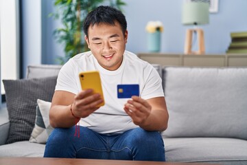 Young chinese man using smartphone and credit card sitting on sofa at home