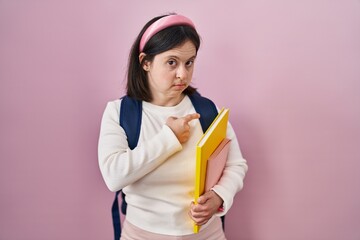 Woman with down syndrome wearing student backpack and holding books pointing with hand finger to the side showing advertisement, serious and calm face