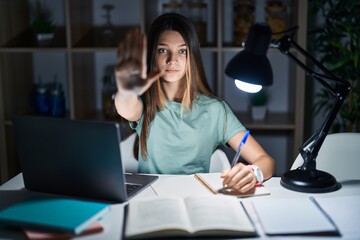 Teenager girl doing homework at home late at night doing stop sing with palm of the hand. warning expression with negative and serious gesture on the face.