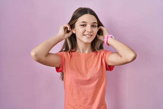 Teenager girl standing over pink background smiling pulling ears with fingers, funny gesture. audition problem