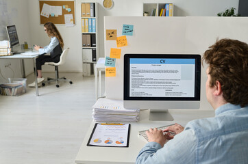 Rear view of young businessman typing resume on computer while sitting at his workplace in office