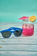 Refreshing summer drink with lots of ice an umbrella and a slice of lemon and blue sunglasses on a wooden table and a greenish background.