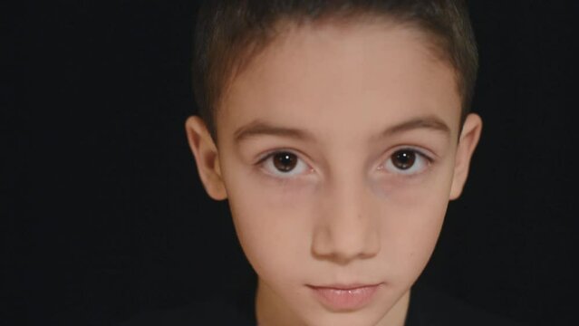 Portrait of young boy raising his head and looking directly to the camera with serious expression. Black background. Childhood and children life. Innocence and age concept.