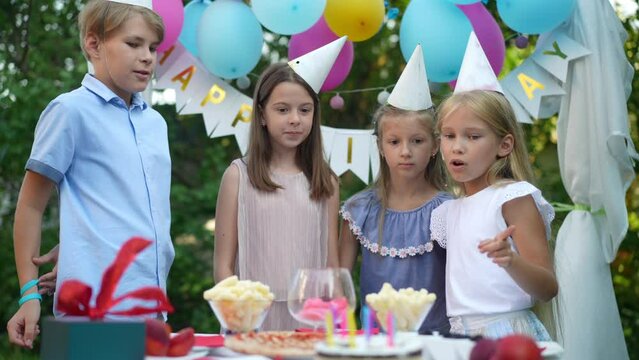 Four cute relaxed kids talking as female hands placing birthday cake on table in slow motion. Excited happy Caucasian boy and girls admiring sweet tasty dessert gesturing thumbs up smiling