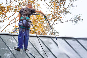 Man, cleaner with high pressure water sprayer wash steel dirty roof.Worker in waterproof uniform...