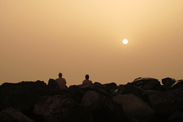 Two lonely figures watch the sunset sitting on the rocks, rear view. Summer holiday.