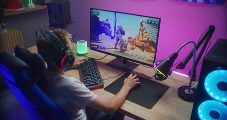 Young gamer plays third person shooter on personal computer. Schoolboy in headphones enjoys online video game at home. Desk illuminated by RGB LED strip light. Concept of gaming at home. Back view. - obrazy, fototapety, plakaty